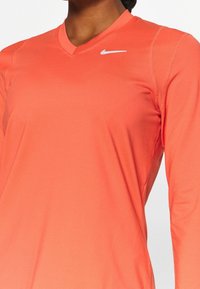Eine Frau trägt ein korallenfarbenes langärmliges Sportshirt mit einem kleinen weißen Nike-Logo auf der oberen Brust.