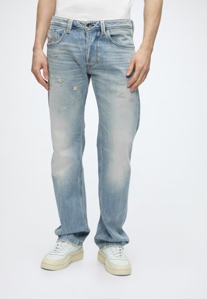 Homme portant un jean délavé bleu clair et des baskets blanches, debout devant un fond blanc uni.