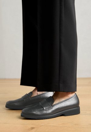 Mocassins en cuir noir portés avec un pantalon noir à jambes larges sur un sol en bois clair, montrant les détails latéraux et avant de la chaussure.