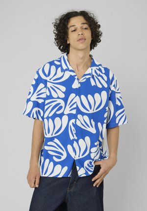 Jeune homme aux cheveux bouclés portant une chemise à manches courtes bleu et blanc avec des motifs abstraits de feuilles, debout devant un fond uni.