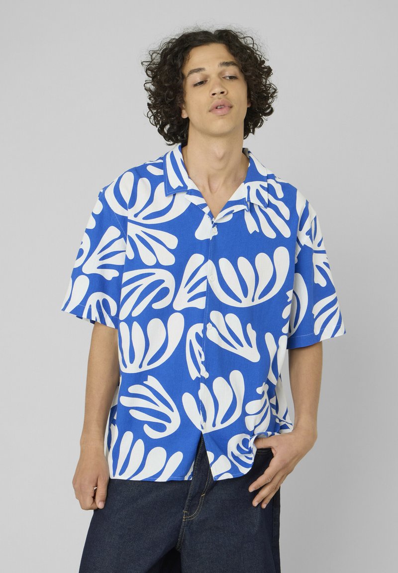 Jeune homme aux cheveux bouclés portant une chemise à manches courtes bleu et blanc avec des motifs abstraits de feuilles, debout devant un fond uni.
