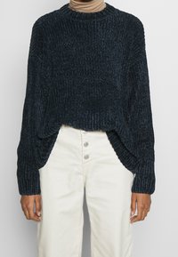 Ciemnogranatowy prążkowany sweter z okrągłym dekoltem, opuszczonymi ramionami i bocznymi rozcięciami, w połączeniu z kremowymi, wysokim stanem spodniami.