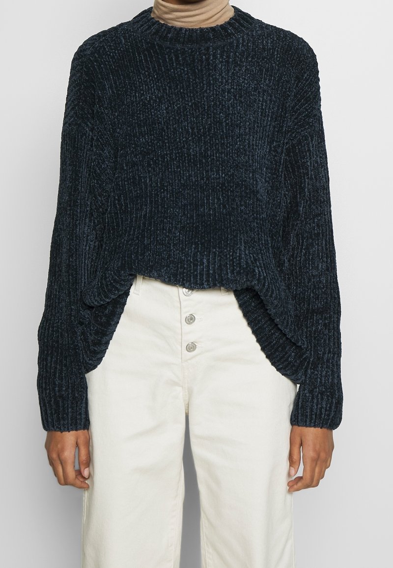 Ciemnogranatowy prążkowany sweter z okrągłym dekoltem, opuszczonymi ramionami i bocznymi rozcięciami, w połączeniu z kremowymi, wysokim stanem spodniami.