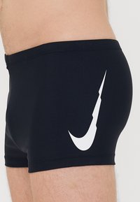 Fekete fürdőnadrág, rugalmas anyagból készült, amelynek oldalán egy kiemelkedő fehér Nike logó található. A dizájn tiszta derékrésszel rendelkezik.