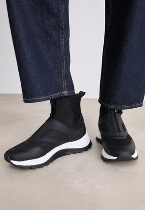 Persona che indossa sneaker alte nere con suole bianche e pantaloni di denim blu scuro, in piedi su un pavimento chiaro.