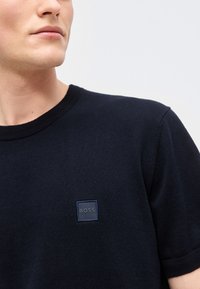 Νεαρό άτομο που φοράει μαύρο κοντομάνικο πουκάμισο με ένα μικρό τετράγωνο λογότυπο "BOSS" στο στήθος, κοιτάζοντας ελαφρώς προς τα αριστερά.