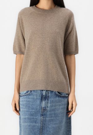 Mujer vistiendo un suéter beige de punto de manga corta combinado con una falda de mezclilla azul hasta la rodilla, de pie sobre un fondo blanco liso.