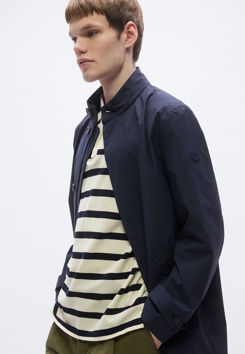 Navy lightweight jacket with a stand collar, paired with a cream and navy striped polo. Dark trousers and subtle button accents visible.