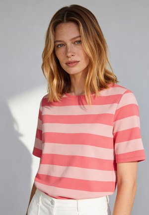 Frau mit schulterlangem blondem Haar trägt ein kurzärmeliges Shirt mit pinken und korallenfarbenen Streifen sowie eine weiße Hose vor einem schlichten Hintergrund.