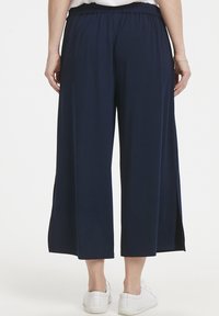 Pantaloni a gamba larga blu navy con una vestibilità comoda, cintura elastica e spacchi laterali, abbinati a sneaker bianche. Texture del tessuto liscia.
