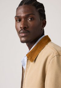 Μπεζ μπουφάν με γιακά μουσταρδί από κορντόρι, κλείσιμο με φερμουάρ, φορεμένο πάνω από ένα μπλε και λευκό πουκάμισο με καρό, που αναδεικνύει ομαλές υφές και μοντέρνο σχέδιο.