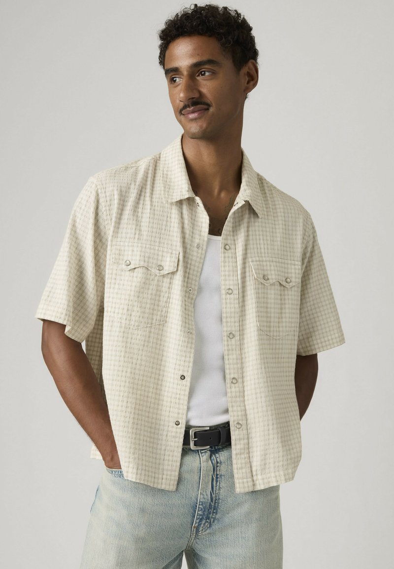 Hombre con cabello corto y rizado que lleva una camisa de manga corta beige a cuadros sobre una camiseta blanca sin mangas y vaqueros azul claro con cinturón negro.