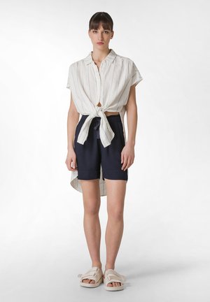 Hvid stribet bluse bundet om taljen, parret med navy shorts og beige sandaler. Blusen har korte ærmer og en afslappet pasform.