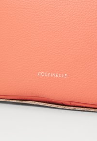 Portafoglio in pelle corallo con finitura testurizzata, con il nome del marchio "Coccinelle" in lettering bianco a rilievo. I bordi presentano cuciture ordinate.