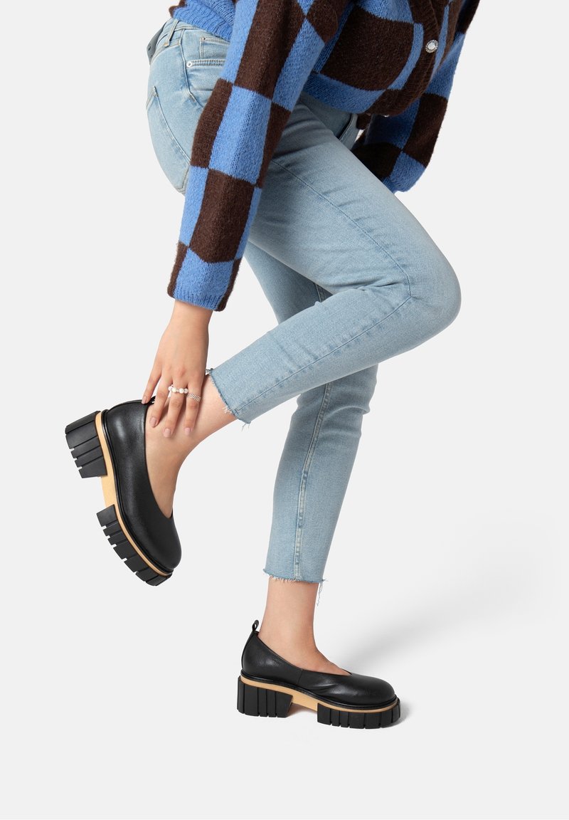 Schwarze Leder-Plateauschuhe mit einem klobigen Holzabsatz, runder Spitze und einer Schlaufe an der Rückseite, kombiniert mit hellblauen Skinny-Jeans.