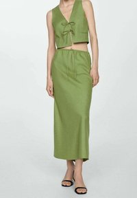 Frau, die ein ärmelloses, olivgrünes Croptop mit vorderen Bindebändern und einen passenden, hochgeschnittenen Midirock trägt, kombiniert mit schwarzen offenen Sandalen.