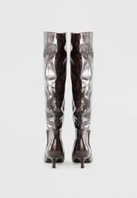 Botas brillantes de color marrón oscuro hasta la rodilla con textura de cocodrilo y pequeños tacones de gato, vistas desde atrás sobre un fondo blanco.