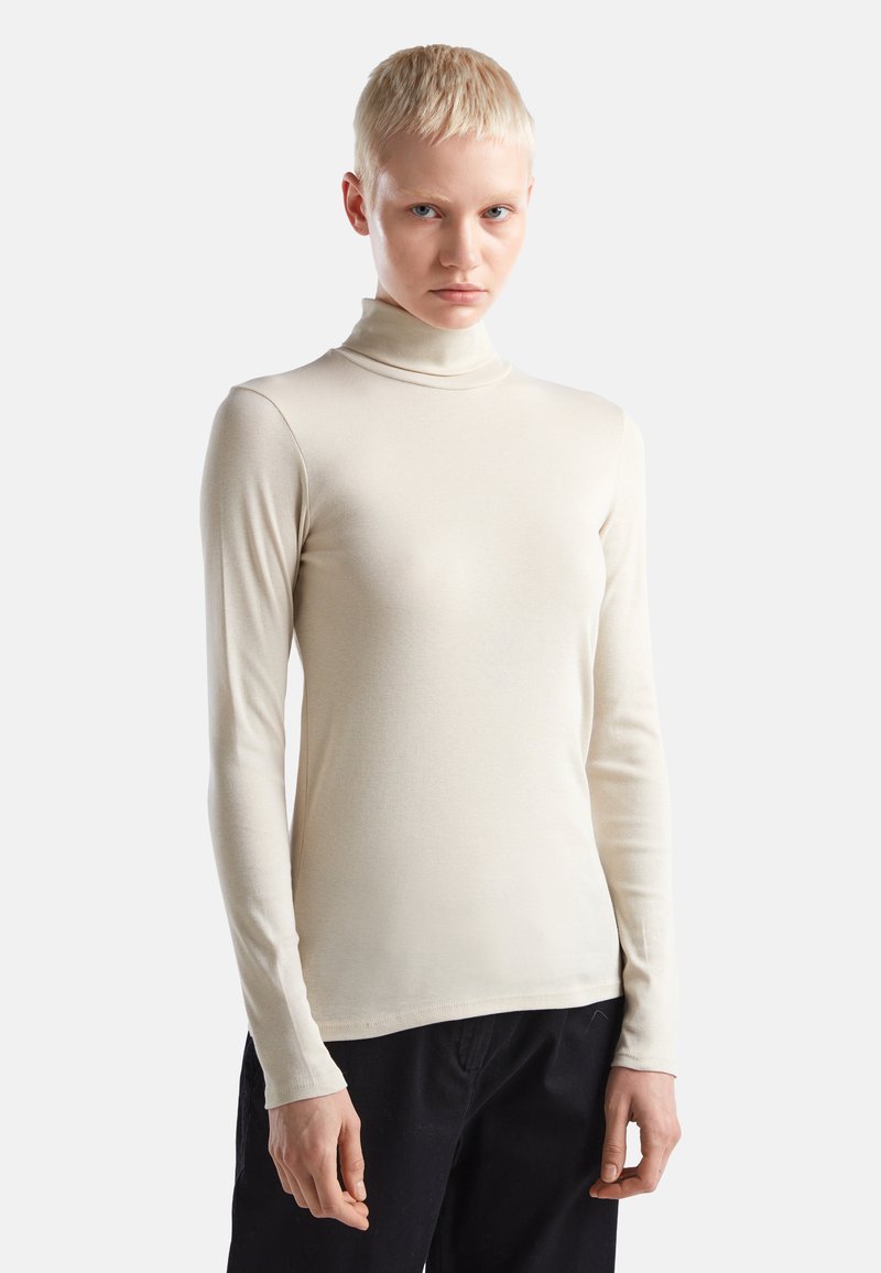 Persona con capelli biondi corti che indossa un dolcevita beige a maniche lunghe e pantaloni neri su uno sfondo bianco uniforme.