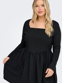 Blond kvinne iført en langermet svart kjole med firkantet utringning, som holder kjoleskjørtet med én hånd.