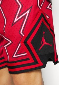 Röda basketshorts med svarta detaljer, med ett nätstruktur, vågmönster och en Jumpman-logotyp på en kontrasterande svart panel.