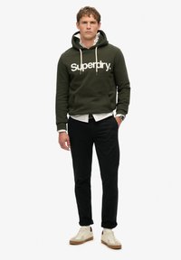 Felpa con cappuccio verde scuro con logo bianco "Superdry", tasca frontale, abbinata a pantaloni neri e sneakers off-white con dettagli marroni.