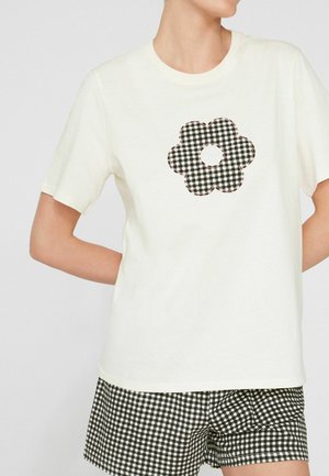 Person trägt ein weißes T-Shirt mit einem schwarz-weiß karierten Blumenmuster, kombiniert mit schwarz-weiß karierten Shorts.
