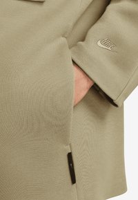 Maslinasto zelena Nike jakna s teksturiranim materijalom, koja ima bočnu džepu i mali izvezeni logo na rukavu.