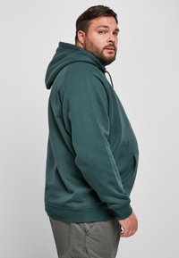 Sudadera con capucha en un tono verde azulado oscuro, confeccionada con una tela suave, que incluye un bolsillo frontal y puños acanalados, con un corte holgado y capucha ajustable con cordón.