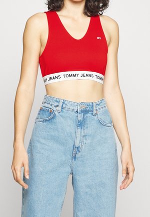 Kvinde iført en rød crop top med "Tommy Jeans" logo-bånd og lyseblå højtaljede denimjeans, stående mod en ensfarvet baggrund.