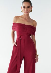 Červený overal s odhalenými ramenami z textúrovaného materiálu, so zúženým pásom opatreným opaskom so zlatou prackou a bočnými vreckami.