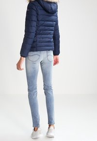 Granatowa kurtka puchowa z kapturem wykończonym futrem, zestawiona z jasnoniebieskimi obcisłymi dżinsami i białymi sneakersami. Posiada pikowaną strukturę i dopasowany krój.