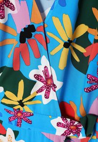 Tissu bleu vif avec de grandes fleurs abstraites en rouge, jaune, rose, blanc et vert, avec des centres de fleurs noirs et orange et des coutures visibles.