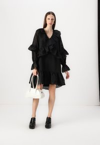 Svart kjole med volanger på ermer og kant, med et teksturert mønster; kombinert med en hvit håndveske og svarte sandaletter.