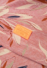 Textiel in een koraal- en crèmekleurenschema met bladvormen. Beschikt over een textuur patch in oranje met reliëflettering.