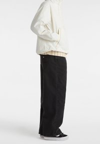 Vit lättviktsjacka med fickor, lager på lager över en beige mönstrad skjorta, kombinerad med lösa svarta jeans och svarta sneakers med vita sulor.