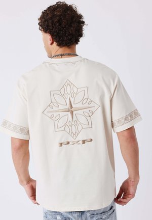 Junger Mann mit lockigem Haar trägt ein weißes T-Shirt mit beigem, geometrischem Stern-Design und dem Schriftzug "PXP" auf dem Rücken, hellblaue Jeans.