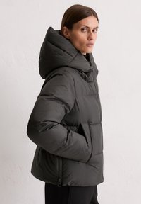 Vrouw in een zwarte pufferjack met grote capuchon, handen in de zakken, kijkt opzij tegen een effen lichte achtergrond.