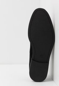 Semelle de chaussure noire avec une surface lisse, légèrement texturée et une petite indication de taille près du talon, reposant contre un coin blanc.