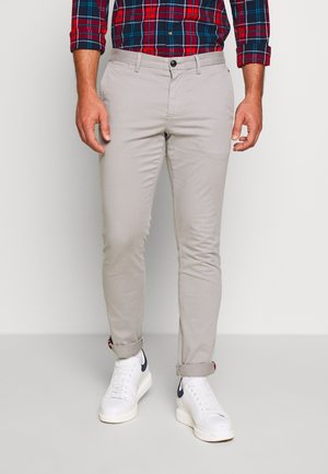 Mann trägt hellbeige Slim-Fit-Hose, rot-blau kariertes Hemd und weiße Sneakers, steht vor einem einfarbigen hellen Hintergrund.