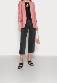 Różowa marynarka, czarny t-shirt z logo, czarne, wysokie, skrócone dżinsy oraz czarne sandały na obcasie. Mała różowa torebka dodaje kontrastu.