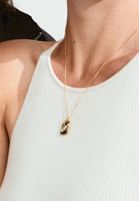Collier pendentif en or avec un charme rectangulaire texturé comportant de petites pierres incrustées, porté sur un haut blanc à côtes.