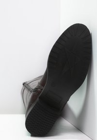 Botte marron foncé avec une tige en cuir lisse, fermeture éclair, et une semelle en caoutchouc texturée présentant un motif en zigzag et un logo.