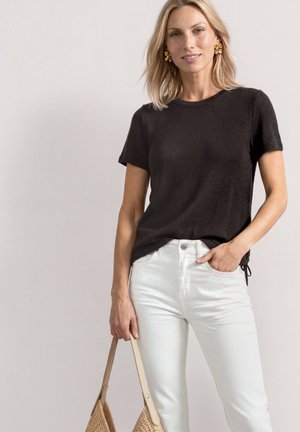 Blondynka ubrana w czarny top z krótkim rękawem i białe spodnie, trzymająca beżową plecioną torebkę z jasnobrązowymi skórzanymi paskami.