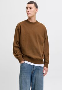 Ung mand iført en brun sweatshirt, hvid undertrøje og løse blå jeans med den ene hånd i lommen, kigger til siden.