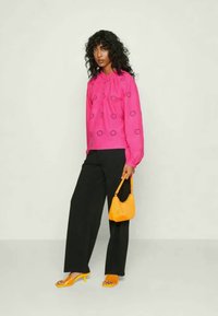 Blusa rosa com enfeites de ilhós e decote redondo, calças largas pretas e acessórios laranja, incluindo saltos e uma carteira.