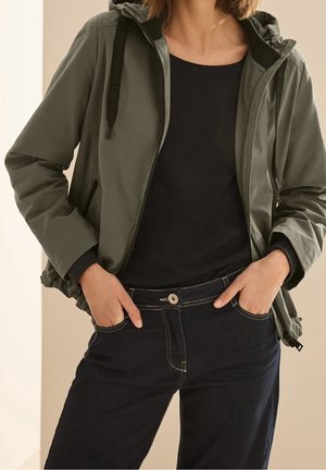 Vrouw met een open-rits olijfgroene jas met capuchon, zwart shirt en donkerblauwe spijkerbroek, met haar handen in de zakken.