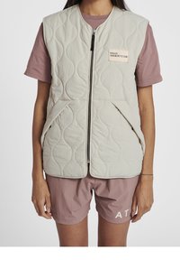 Quilted beige vest with a front zipper, featuring a pocket with a label and curved stitching patterns, worn over a pink shirt and shorts.