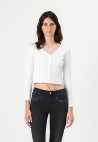 Cárdigan corto de canalé blanco con mangas largas y cierre de botones, combinado con jeans negros que cuentan con adornos decorativos en los bolsillos.