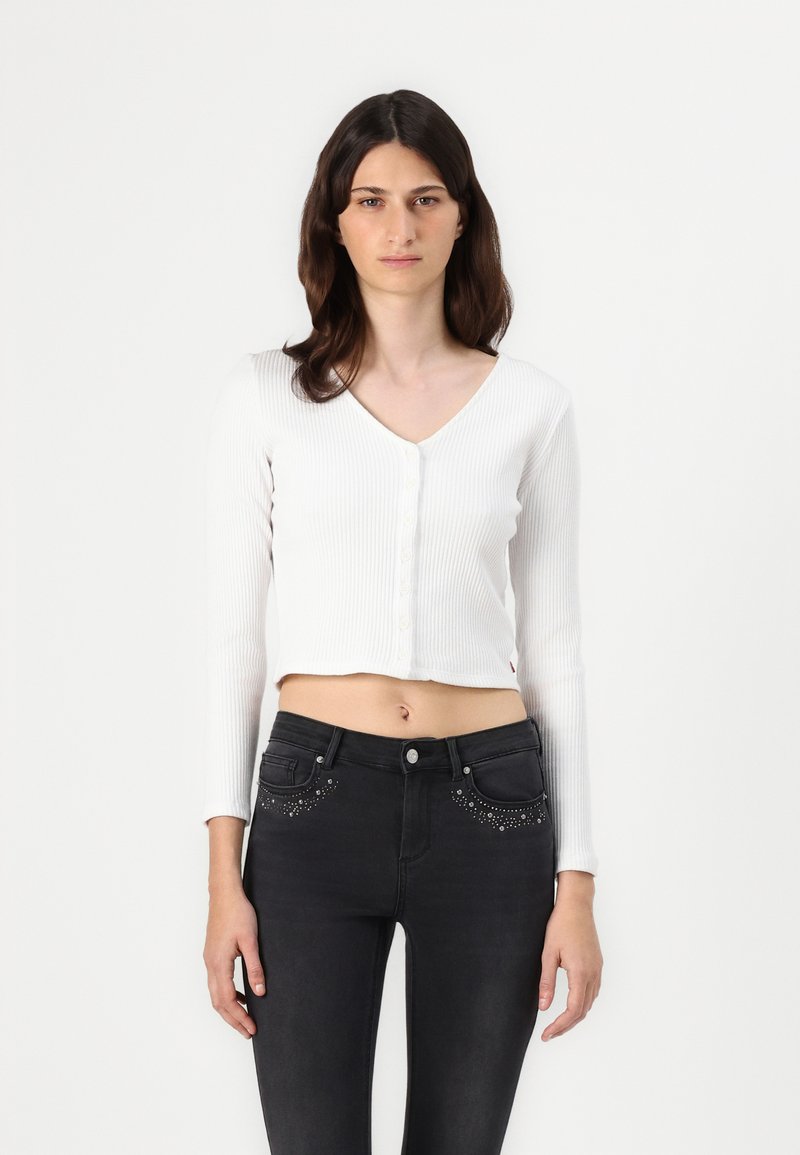 Cárdigan corto de canalé blanco con mangas largas y cierre de botones, combinado con jeans negros que cuentan con adornos decorativos en los bolsillos.