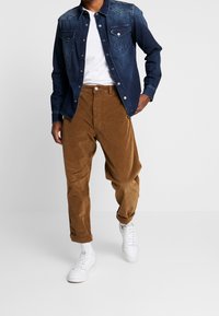 Camicia di jeans con gradiente dal blu chiaro al blu scuro, due tasche sul petto; pantaloni in corduroy marroni; sneakers bianche; vestibilità rilassata, orli arrotolati sui pantaloni.